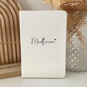 Op de afbeelding: Een wit canvas notitieboekje met de naam "Madeline" geschreven in zwarte cursieve letters en een klein zwart hartje.