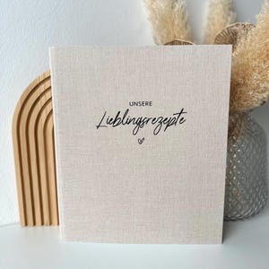 Op de afbeelding: Een beige receptenboek met linnen omslag met de woorden "UNSERE Lieblingsrezepte" in zwart schrift. Onder de titel staat een klein hartje. Het boek wordt getoond met decoratief pampasgras en een houten boog.