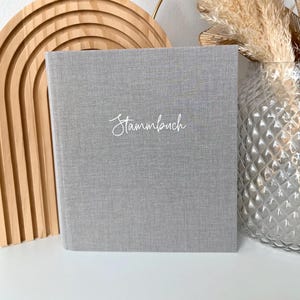 Könnte beinhalten: Ein graues Hardcover-Buch mit dem Wort "Stammbuch" in weißer Schrift. Das Buch steht vor einem neutralen Hintergrund mit dekorativen Elementen, darunter ein Holzbogen und Trockenblumen in einer Glasvase.
