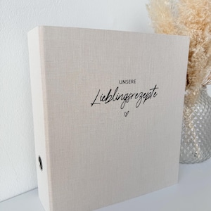 Op de afbeelding: Een beige ordner met de woorden "UNSERE Lieblingsrezepte" in zwart schrift, met een klein hartjessymbool. De ordner staat rechtop, met een zilveren vaas en droogbloemen op de achtergrond. De ordner heeft een ringmechanisme.