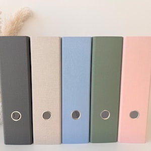 Könnte beinhalten: Eine Reihe von fünf Ordnern in verschiedenen Farben: Dunkelgrau, Beige, Hellblau, Olivgrün und Rosa. Jeder Ordner hat einen goldfarbenen Metallringgriff. Die Ordner sind vertikal auf einer weißen Oberfläche angeordnet.