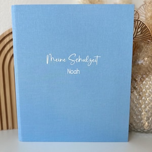 Könnte beinhalten: Ein hellblaues Hardcover-Buch mit dem Text "Meine Schulzeit Noah" in weißer Schrift. Das Buch hat einen strukturierten Einband und steht aufrecht. Der Hintergrund enthält dekorative Elemente.