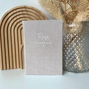 Könnte beinhalten: Ein hellgraues Notizbuch mit den Wörtern "Reise" und "Tagebuch" auf dem Einband. Das Notizbuch steht vor einem Holzrahmen und einer Vase mit dekorativen Elementen.