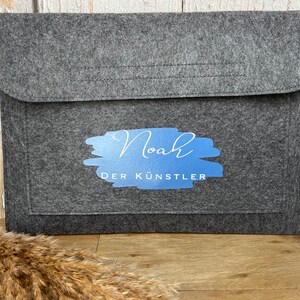 Könnte beinhalten: Eine graue Filz-Laptoptasche mit Klappenverschluss. Die Tasche hat ein blau-weißes Design mit dem Namen "Noah" und den Worten "DER KÜNSTLER". Sie dient zum Schutz eines Laptops oder Tablets.