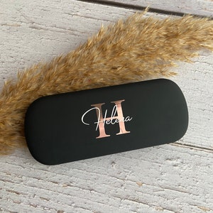 Könnte beinhalten: Schwarze Brillenetui mit einem roségoldenen Initial "H" und dem Namen "Helena" in Kursivschrift.