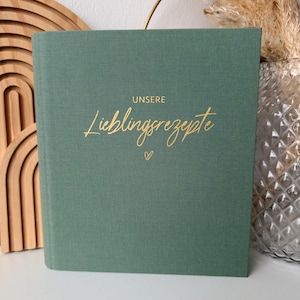 Könnte beinhalten: Ein salbeigrünes, stoffgebundenes Rezeptbuch mit dem goldfarbenen Schriftzug "UNSERE Lieblingsrezepte" und einem kleinen Herzsymbol in der Mitte des Einbands. Das Buch hat eine klare, rechteckige Form mit einer strukturierten, matten Oberfläche.