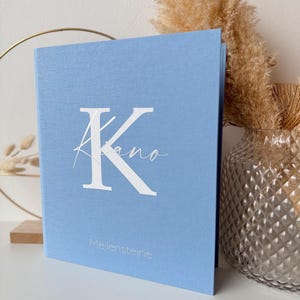 May include: A light blue book with the letter "K" and the name "Kiano" in white script. The word "Meilensteine" is printed in a smaller font below. The book is standing upright, with a neutral background.