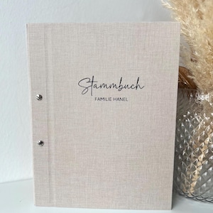 Op de afbeelding: Een lichtbeige stoffen Stammbuch, of familieboek, met de woorden "Stammbuch FAMILIE HANEL" in zwart schrift. Het boek heeft drie zilveren bindringen op de rug.