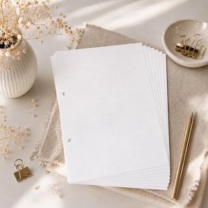 Könnte beinhalten: Ein Stapel weißer, rechteckiger Papierbögen mit zwei kreisförmigen Löchern am linken Rand. Das Papier liegt auf einem beigen Stofftuch neben einem goldfarbenen Stift, einer kleinen Keramikschale mit goldenen Büroklammern und einer Vase.