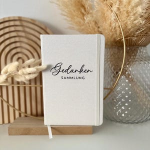 Könnte beinhalten: Ein weißes Notizbuch mit einer schwarzen Aufschrift, die "Gedanken Sammlung" lautet. Das Notizbuch liegt auf einer Holzoberfläche mit einem goldenen Metallring und einer Vase mit getrocknetem Pampasgras im Hintergrund.
