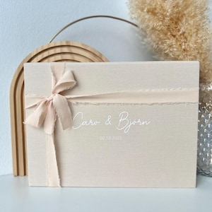 May include: A beige rectangular box tied with a light pink ribbon and bow. The box has the names "Caro & Bjorn" and the date "02.10.2025" printed on the front. The box is next to a wooden arch and a vase with dried flowers.