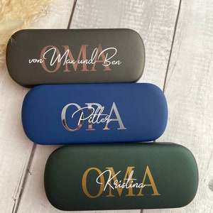 Könnte beinhalten: Drei personalisierte Brillenetuis. Das obere Etui ist schwarz mit roségoldenen Lettern, auf denen "von Max und Ben OMA" steht. Das mittlere Etui ist blau mit silbernen Lettern, auf denen "OPA Pitter" steht. Das untere Etui ist grün mit goldenen Lettern, auf denen "OMA Kristina" steht.
