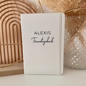Op de afbeelding: Wit linnen gebonden dagboek met de tekst "ALEXIS Traumtagebuch" in zwart gedrukt.
