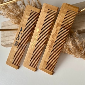 Könnte beinhalten: Drei Holz-Kämme mit personalisierten Gravuren. Die Kämme sind aus natürlichem Bambus und haben eine hellbraune Farbe. Die Gravuren sind in goldener und schwarzer Schrift. Die Kämme sind in einer Reihe angeordnet, wobei der gravierte Text zum Betrachter zeigt. Der Text auf den Kämmen lautet "Opa Gerry", "Amelie" und "Weltbeste Mama".