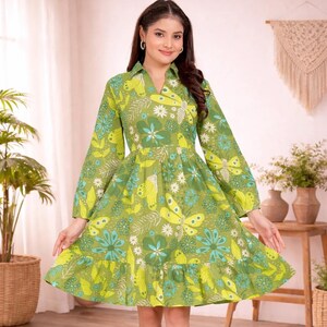Könnte beinhalten: Ein grünes Kleid mit Schmetterlings- und Blumenmuster. Das Kleid hat lange Ärmel, einen V-Ausschnitt und einen Rüschensaum. Das knielange Kleid ist aus leichtem Stoff gefertigt. Geeignet für Freizeitkleidung, es ist ein lebendiges und stilvolles Kleidungsstück.