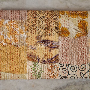 Könnte beinhalten: Ein Patchwork-Textil mit verschiedenen Mustern und Farben. Das Design umfasst Quadrate aus cremefarbenen, goldenen und rosa Stoffen mit floralen, Paisley- und geometrischen Motiven. Die Kanten sind mit rotem und weißem Faden genäht.