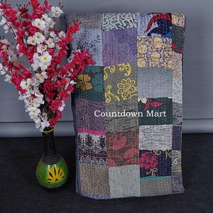 Könnte beinhalten: Ein Patchwork-Quilt mit quadratischen Stoffen in Grau-, Lila-, Grün- und Rosatönen. Der Quilt hat sichtbare Nähte und der Text "Countdown Mart" ist sichtbar.