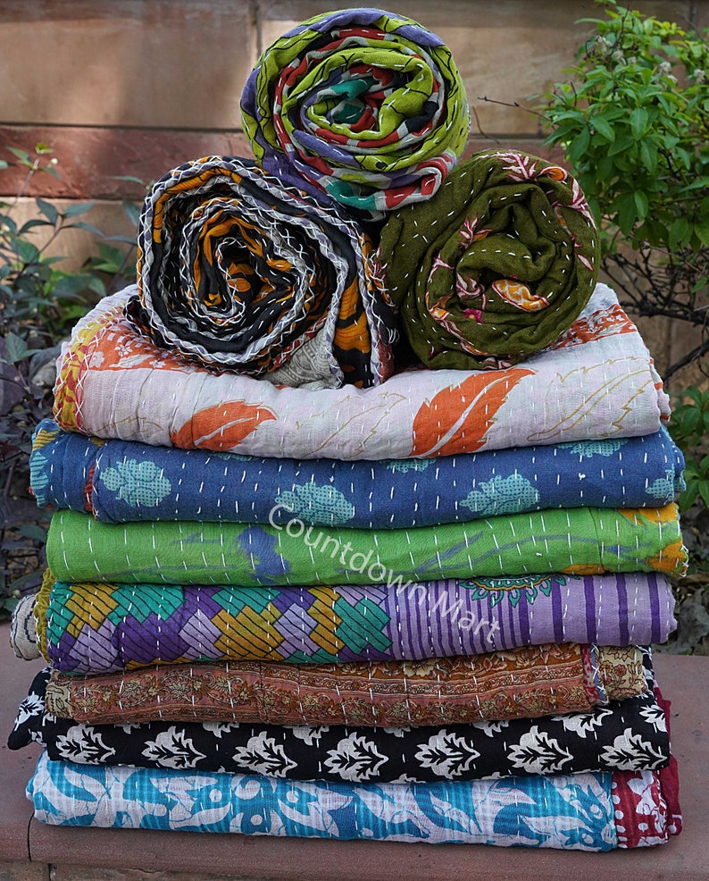 May include: A stack of 11 kantha quilts in various colors and patterns, including floral, geometric, and paisley designs. The quilts are folded and stacked on top of each other, with the top three quilts rolled up.