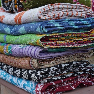May include: A stack of colorful kantha quilts, a traditional Indian textile, with various patterns and stitching. The quilts are folded and stacked on top of each other, creating a visually appealing display.