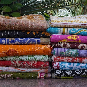 May include: A stack of colorful kantha quilts with various floral and geometric patterns. The quilts are made of recycled saris and are hand-stitched. The quilts are arranged in a pile, with the top quilt folded in half. The quilts are a vibrant mix of colors, including red, orange, yellow, green, blue, and purple.
