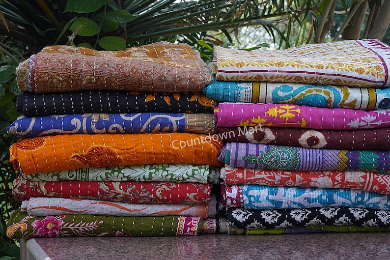 May include: A stack of colorful kantha quilts with various floral and geometric patterns. The quilts are made of recycled saris and are hand-stitched. The quilts are arranged in a pile, with the top quilt folded in half. The quilts are a vibrant mix of colors, including red, orange, yellow, green, blue, and purple.