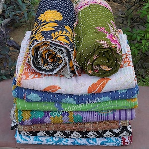 May include: A stack of ten kantha quilts in various colors and patterns, including black, green, blue, orange, and purple. The quilts are folded and stacked on top of each other. The top two quilts are rolled up.