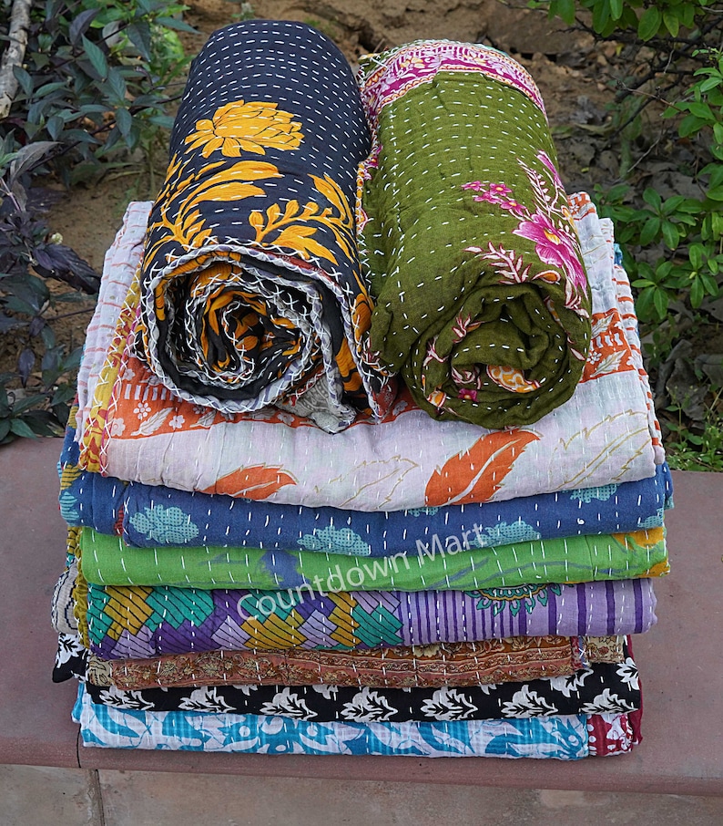 May include: A stack of ten kantha quilts in various colors and patterns, including black, green, blue, orange, and purple. The quilts are folded and stacked on top of each other. The top two quilts are rolled up.