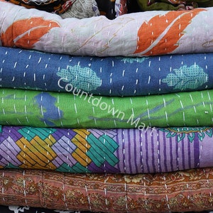 May include: A stack of colorful kantha quilts with various patterns, including floral, geometric, and striped designs. The quilts are made of recycled saris and are hand-stitched with kantha embroidery.