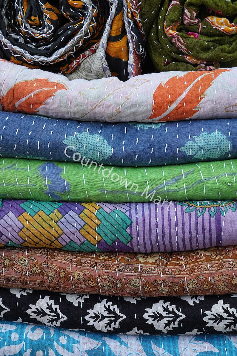May include: A stack of colorful kantha quilts with various patterns, including floral, geometric, and striped designs. The quilts are made of recycled saris and are hand-stitched with kantha embroidery.
