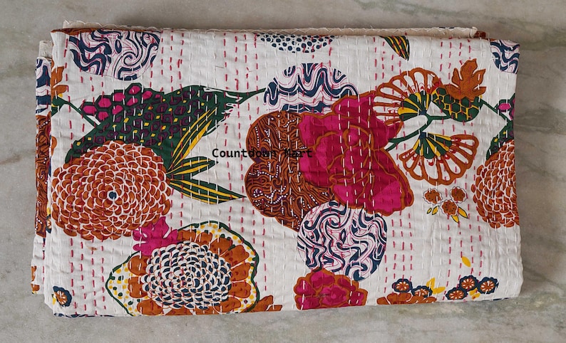 May include: A folded white quilt with a colorful floral pattern. The design includes large flowers in shades of pink, orange, and brown, with green and yellow leaves. The quilt is stitched with red thread.