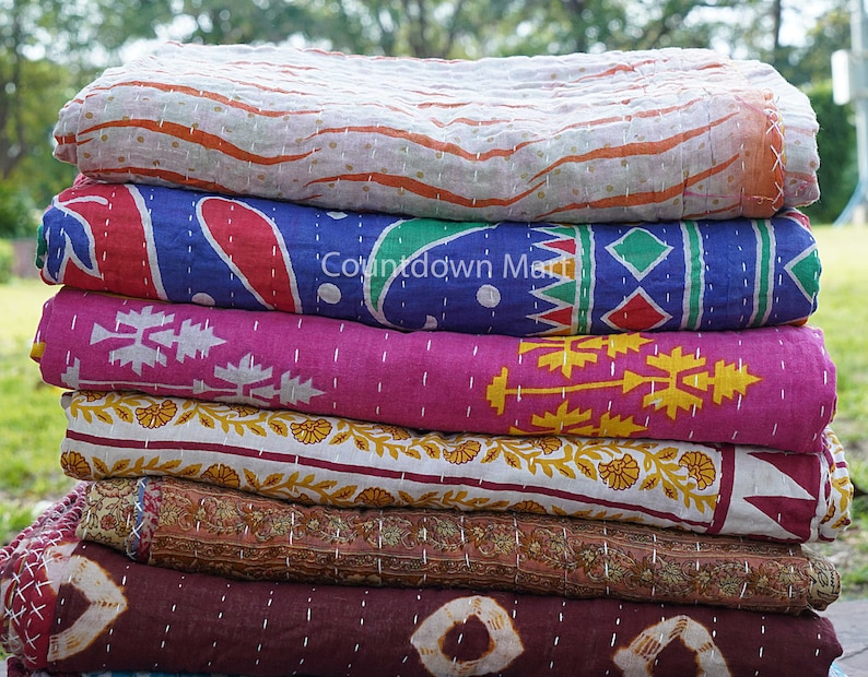 May include: A stack of six colorful kantha quilts with various patterns, including floral, geometric, and paisley designs. The quilts are made of cotton and are hand-stitched. The quilts are folded and stacked on top of each other. The text "Countdown Market" is visible on the second quilt from the top.