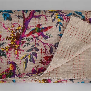 May include: Beige quilt with a colorful floral and bird pattern. The design features vibrant pink, purple, yellow, and blue accents. The quilt is folded, revealing the reverse side with red stitching.