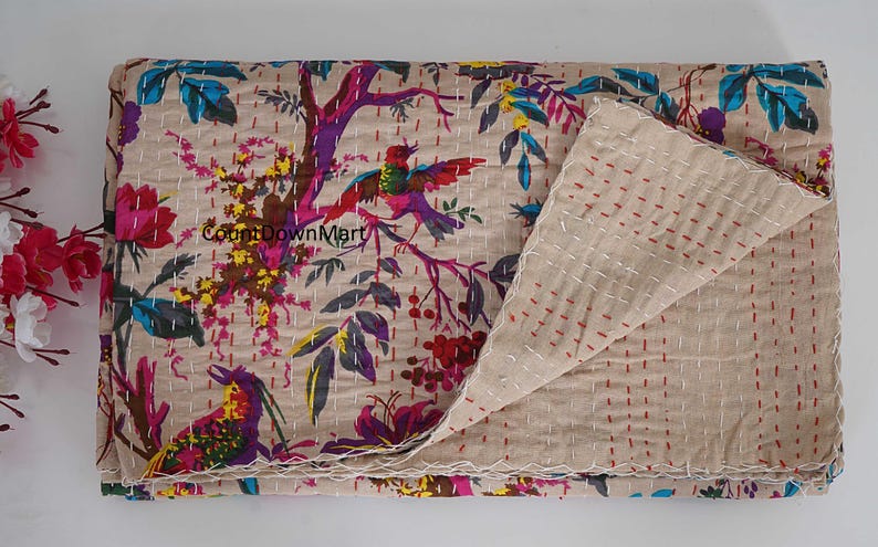 May include: Beige quilt with a colorful floral and bird pattern. The design features vibrant pink, purple, yellow, and blue accents. The quilt is folded, revealing the reverse side with red stitching.