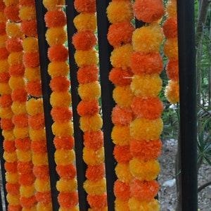 Op de afbeelding: Een guirlande van oranje en gele margrietbloem pompons die aan een zwart metalen hek zijn geregen.