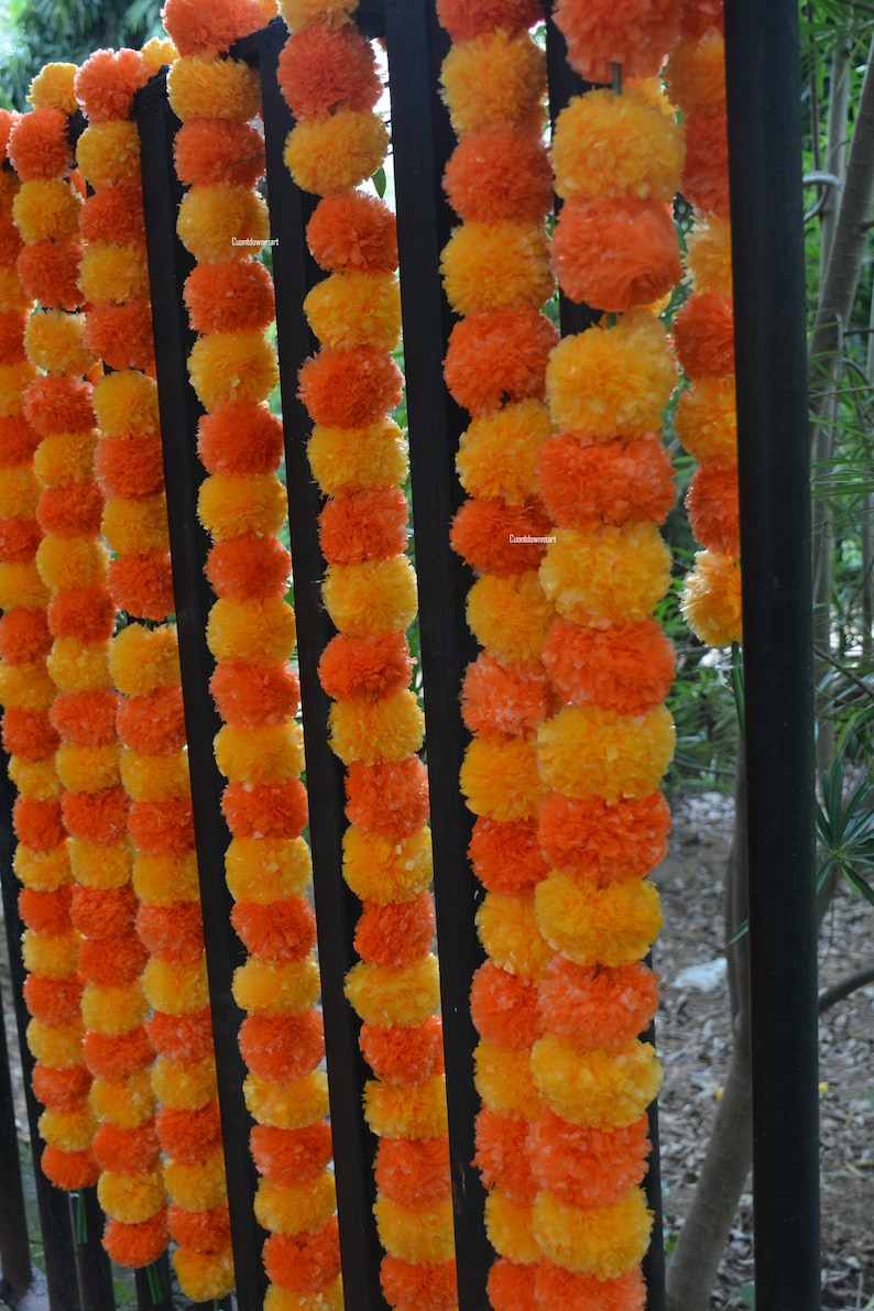 Op de afbeelding: Een guirlande van oranje en gele margrietbloem pompons die aan een zwart metalen hek zijn geregen.