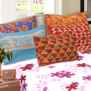 Könnte beinhalten: Vier gemusterte Kissenbezüge in verschiedenen Farben und Designs, darunter Rot, Orange, Blau und Grün, auf einem weißen Bettbezug mit rosafarbenem Blumenmuster angeordnet.