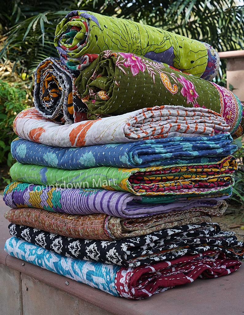 May include: A stack of colorful kantha quilts, a traditional Indian textile, with various floral and geometric patterns. The quilts are rolled up and stacked on top of each other, creating a visually appealing display.