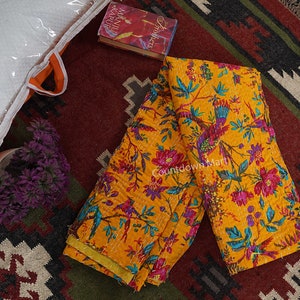 Op de afbeelding: Een gele kantha-deken met een bloemen- en vogelprint. De deken is dubbelgevouwen en ligt op een tapijt met een geometrisch patroon. De deken is gemaakt van katoen en is met de hand gestikt.
