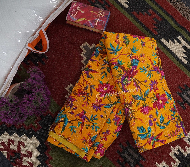 May include: A yellow kantha quilt with a floral and bird print. The quilt is folded in half and is lying on a rug with a geometric pattern. The quilt is made of cotton and is hand-stitched.