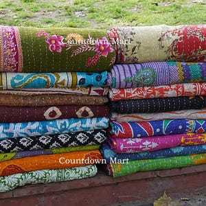 Może przedstawiać: Stos kolorowych, zwiniętych kołder kantha. Kołdry prezentują r&oacute;żnorodne wzory i kolory, w tym kwiatowe, paisley i geometryczne. Tekst "Countdown Mart" jest widoczny na niekt&oacute;rych kołdrach.