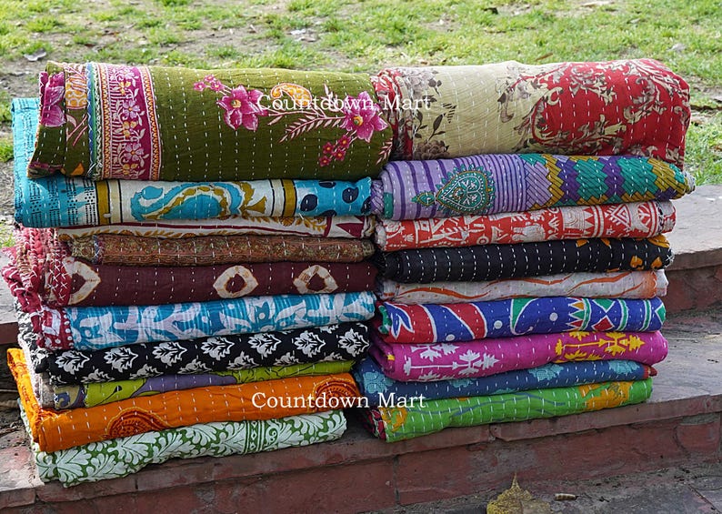 Może przedstawiać: Stos kolorowych, zwiniętych kołder kantha. Kołdry prezentują r&oacute;żnorodne wzory i kolory, w tym kwiatowe, paisley i geometryczne. Tekst "Countdown Mart" jest widoczny na niekt&oacute;rych kołdrach.