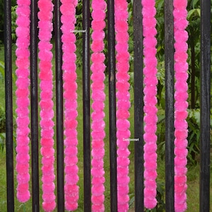 Op de afbeelding: Zeven roze bloemenkransen hangen aan een zwart metalen hek. De kransen zijn gemaakt van pluche pompoenen.