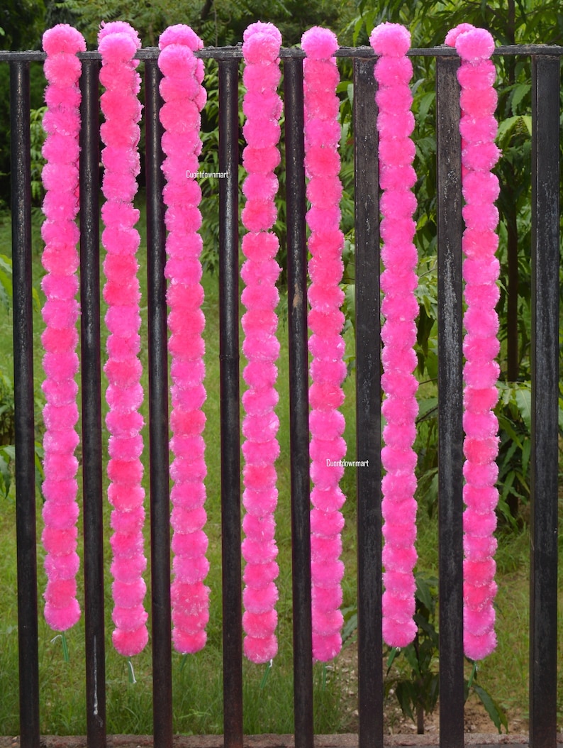 Op de afbeelding: Zeven roze bloemenkransen hangen aan een zwart metalen hek. De kransen zijn gemaakt van pluche pompoenen.