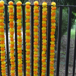 Op de afbeelding: Een reeks gele en oranje margrietbloemenslingers hangt aan een zwart metalen hek. De slingers zijn gemaakt van kunstbloemen en zijn aan elkaar geregen met groen touw.