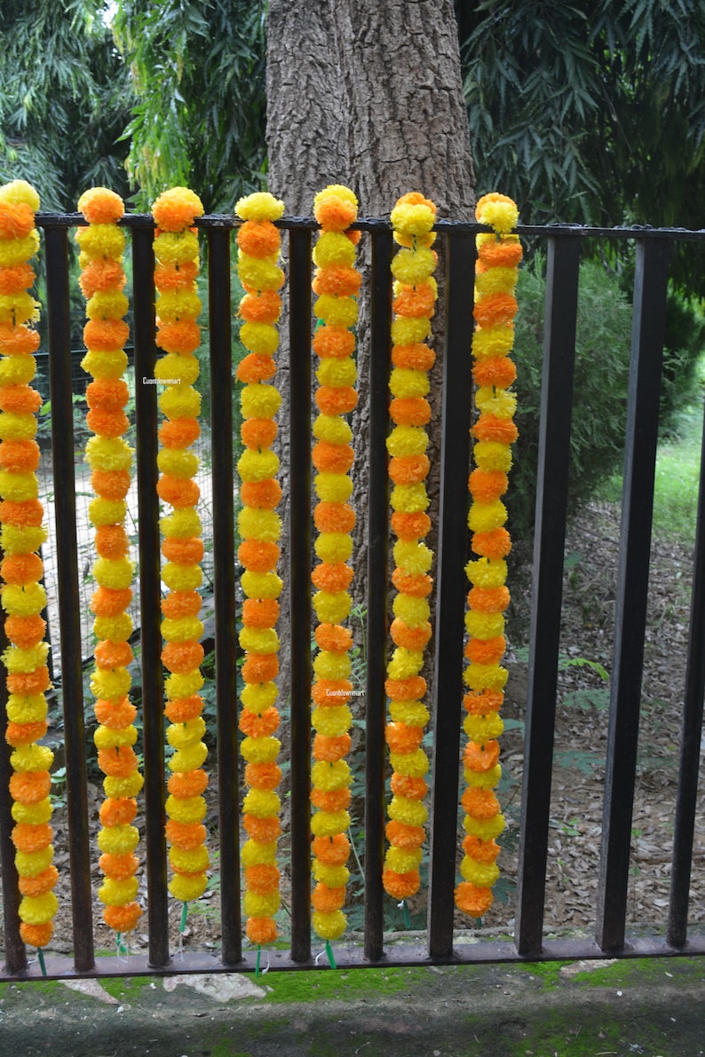 Op de afbeelding: Een reeks gele en oranje margrietbloemenslingers hangt aan een zwart metalen hek. De slingers zijn gemaakt van kunstbloemen en zijn aan elkaar geregen met groen touw.