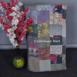 May include: A patchwork quilt with a variety of patterned fabric squares in shades of gray, purple, green, and pink. The quilt has visible stitching and the text "Countdown Mart". A vase with red and white flowers is on the left.