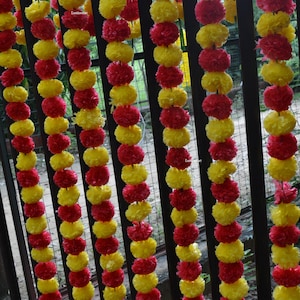 Op de afbeelding: Een guirlande van afwisselend rode en gele bloemen van stof, aan elkaar geregen en hangend aan een zwart metalen hek.
