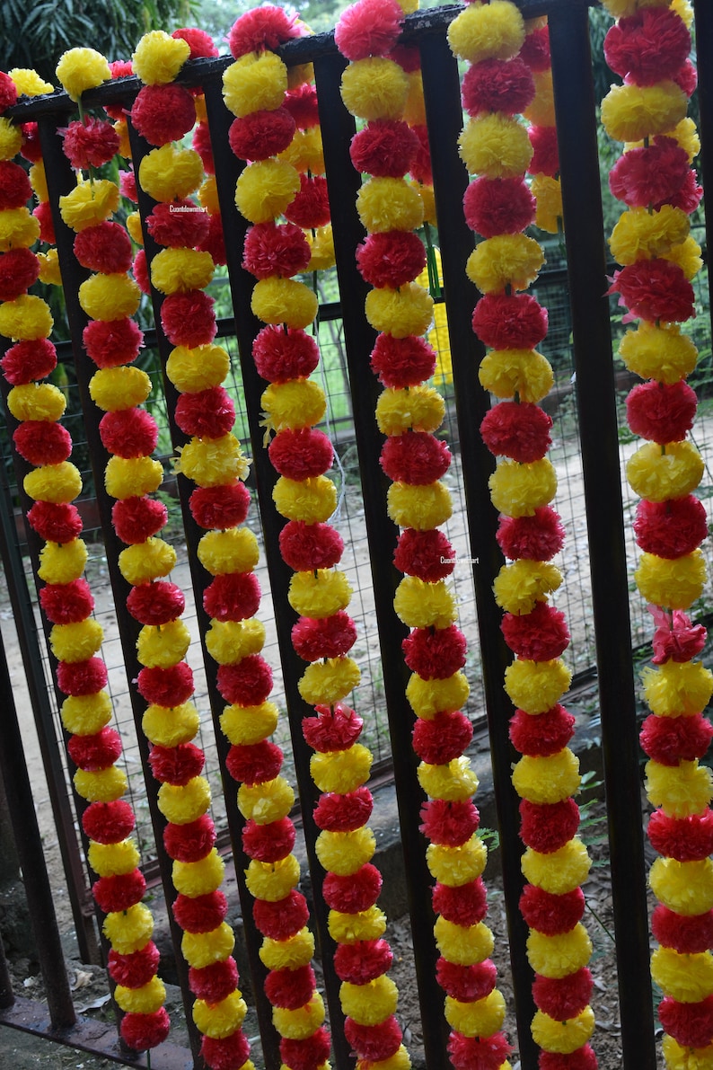 Op de afbeelding: Een guirlande van afwisselend rode en gele bloemen van stof, aan elkaar geregen en hangend aan een zwart metalen hek.