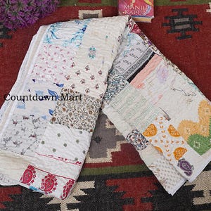 Könnte beinhalten: Eine gefaltete Patchwork-Steppdecke mit verschiedenen floralen und geometrischen Mustern in Creme-, Weiß- und Pastelltönen. Die verschiedenen Abschnitte der Steppdecke zeigen einzigartige Designs. Der Text "Countdown Mart" ist sichtbar.