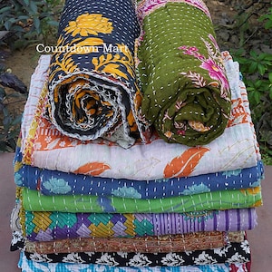May include: A stack of colorful, patterned quilts. Two rolled quilts, one navy with a yellow floral design and one green with pink floral accents, sit atop the stack. The other quilts feature various patterns and colors, including orange, blue, and purple.